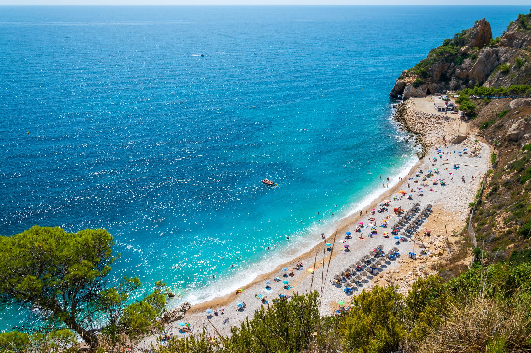 Aerial view of Cala del Moraig in season, swimmers in clear turquoise water off the pale pebble beach